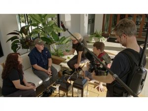 Vier Personen sitzen auf einem Sofa in einer Gesprächssituation. Daneben stehend eine Person mit Mikrofon und Filmkamera.